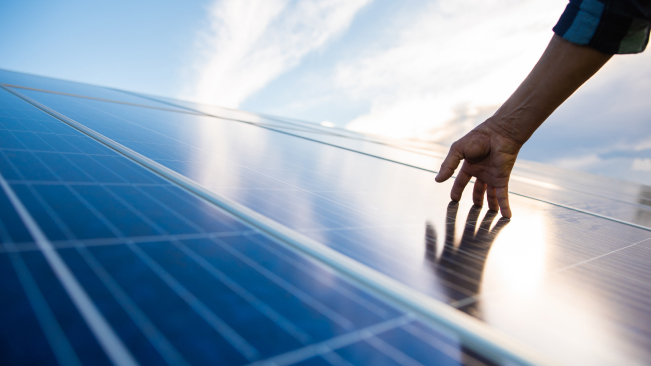 Mão tocando painéis solares sob o sol, simbolizando o uso de energia sustentável e inovação tecnológica.